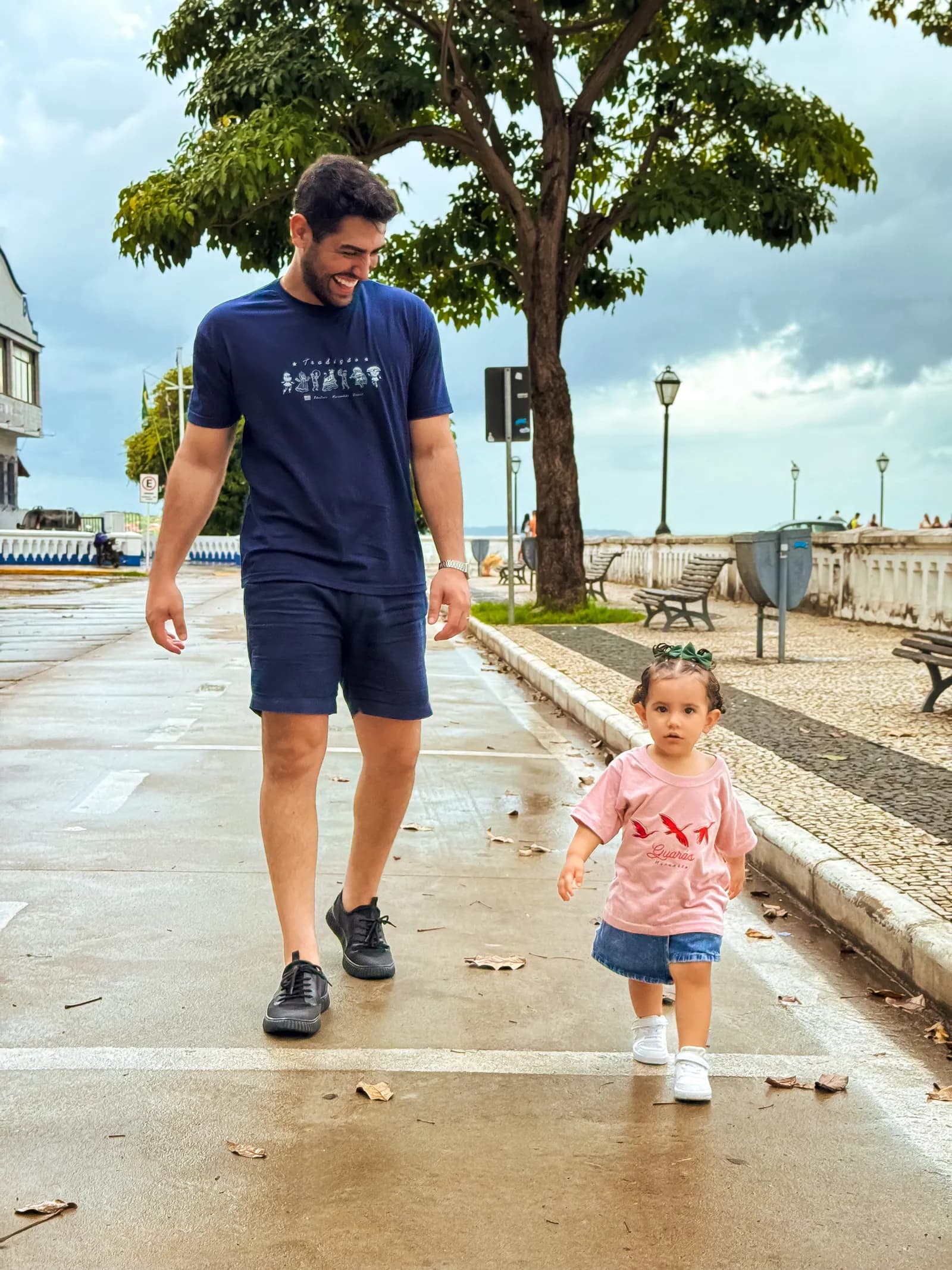Pai e filha caminhando de mãos dadas por uma rua do centro histórico de São Luís, os dois vestindo camisetas da Kambada