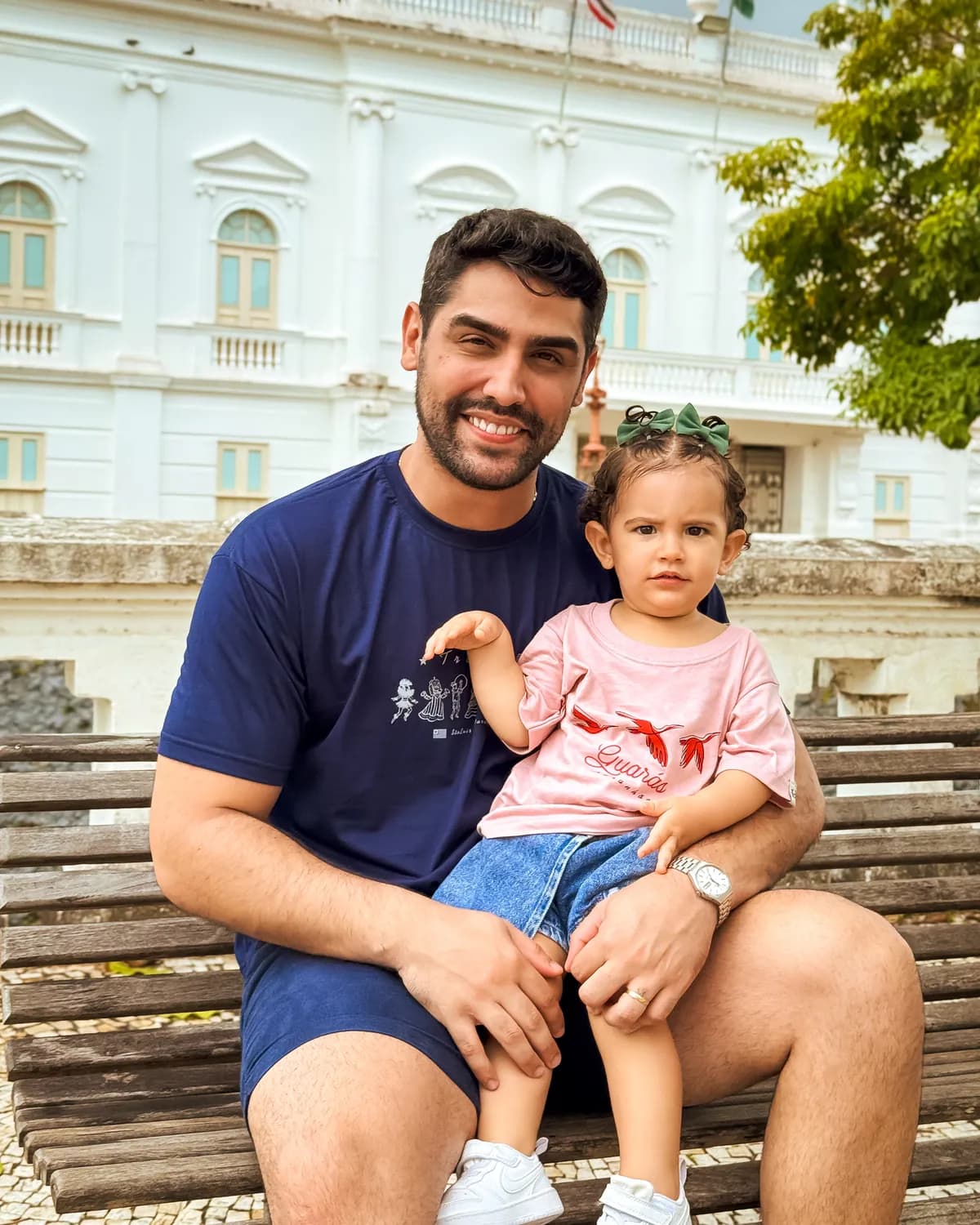 Pai sentado com a filha no colo em frente a um casarão colonial de São Luís, ele de camiseta Tradição e ela de camiseta Guarás