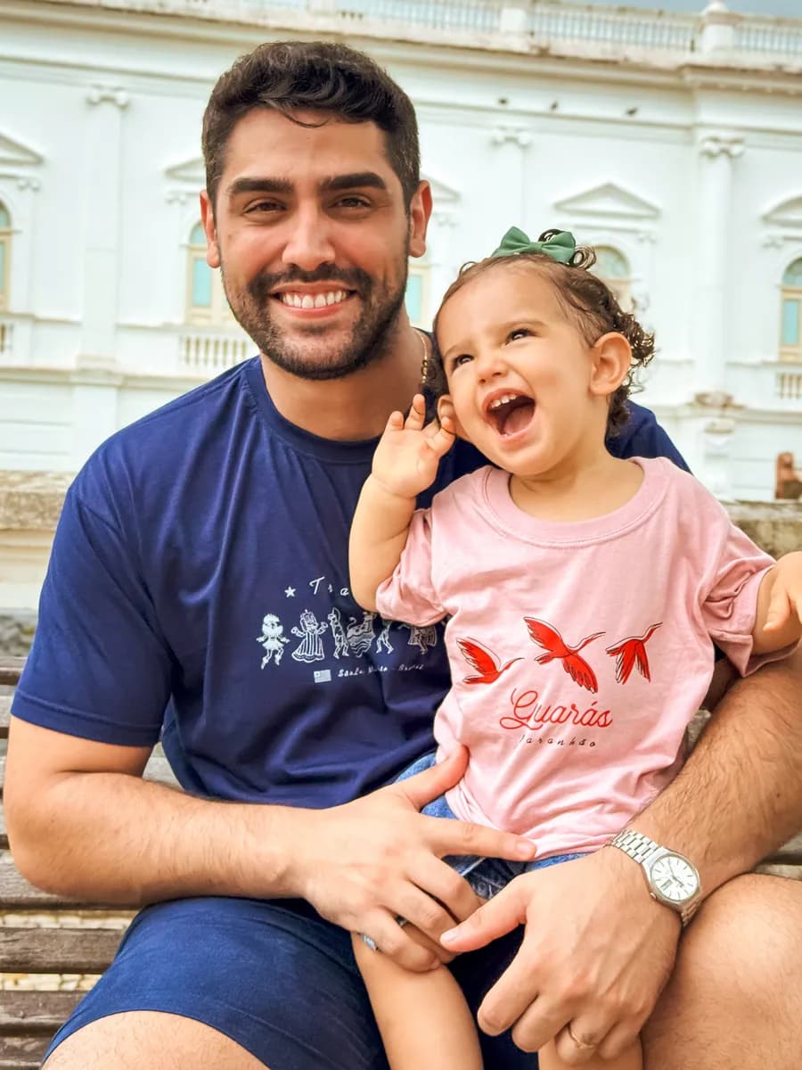 Menina rindo no colo do pai, vestindo a camiseta infantil Guarás, da Kambada